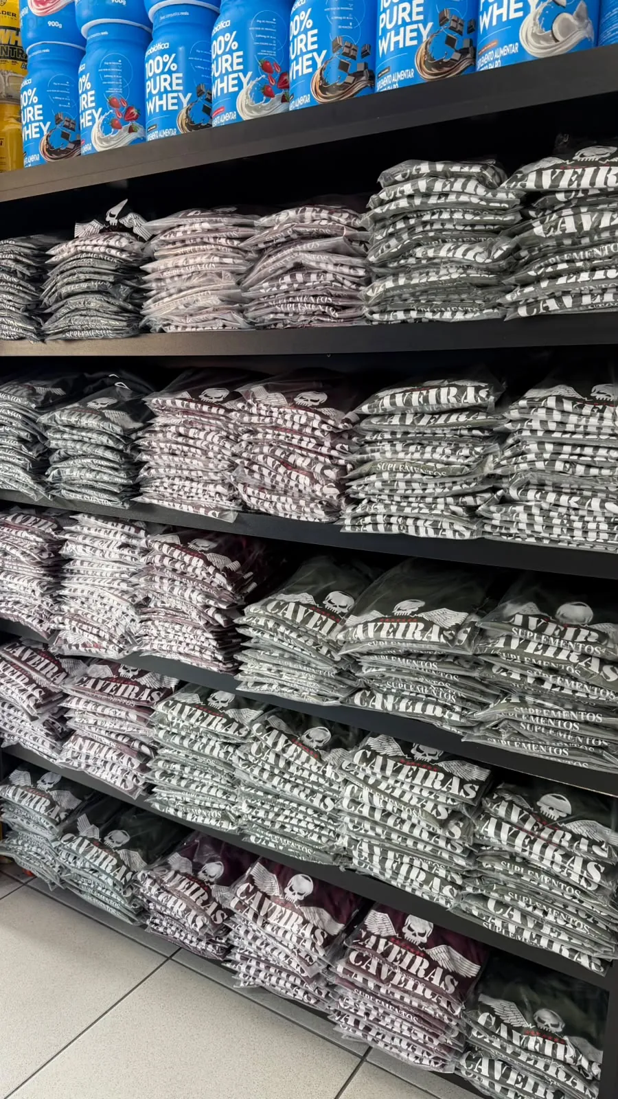 Prateleiras com pilhas de camisetas dobradas com o logo da Caveiras Suplementos, em mais de uma cor