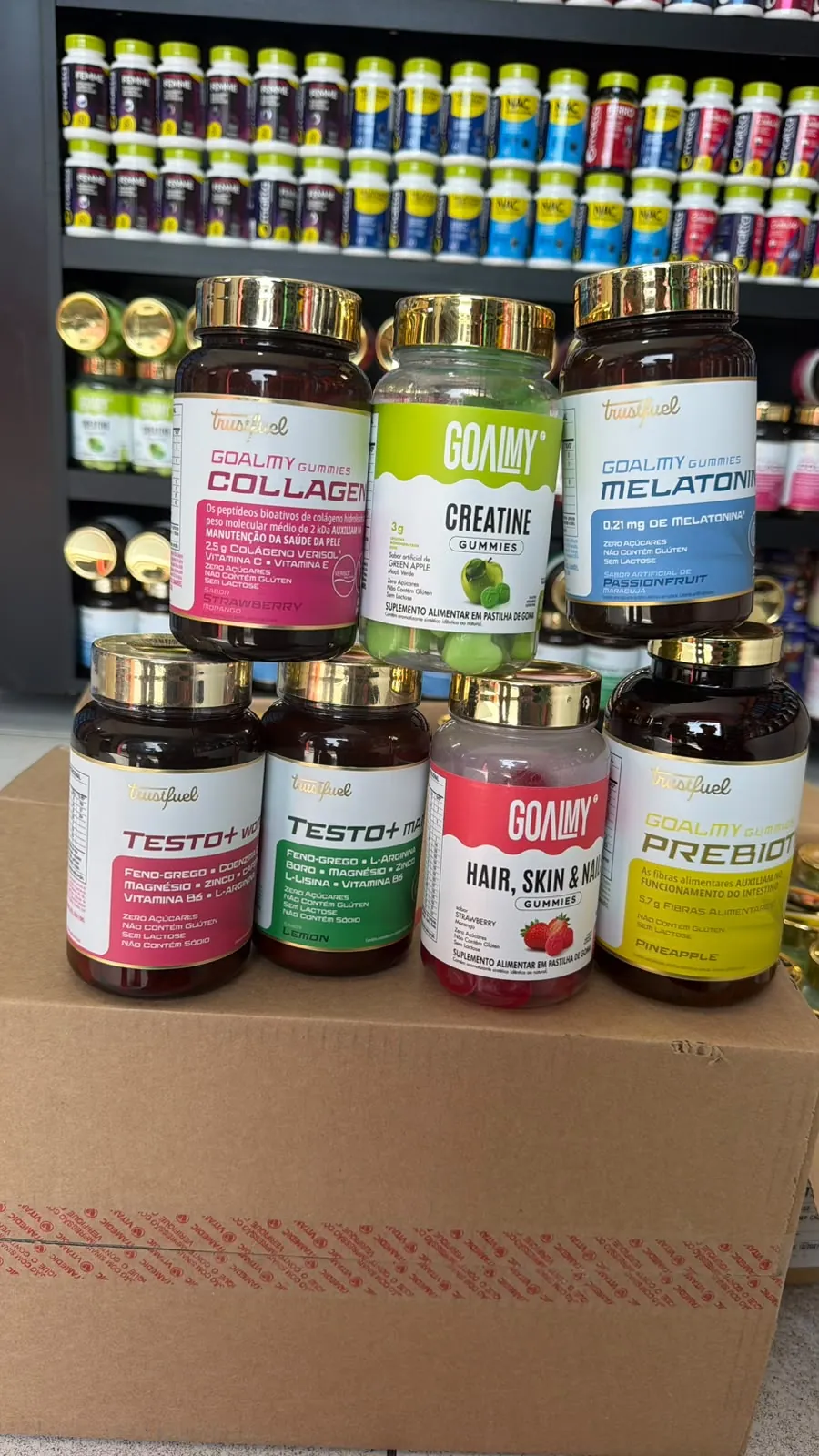 Sete frascos da Trustfuel em cima de uma caixa: gomas Goalmy de colágeno, creatina, melatonina, fibras e cabelo, pele e unhas, e os frascos Testo+ Woman e Testo+ Man